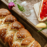 Braided brioche bread with a golden crust, sprinkled with pearl sugar, served on a wooden tray with jam and butter.